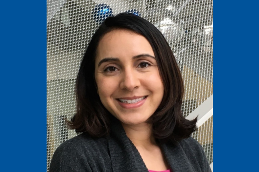 Pictured is Dr. Ophelia Venturelli, PhD, a woman with shoulder-length dark brown hair smiling warmly at the camera. She is wearing a gray sweater over a pink top, standing in front of a light-colored mesh background with soft lighting